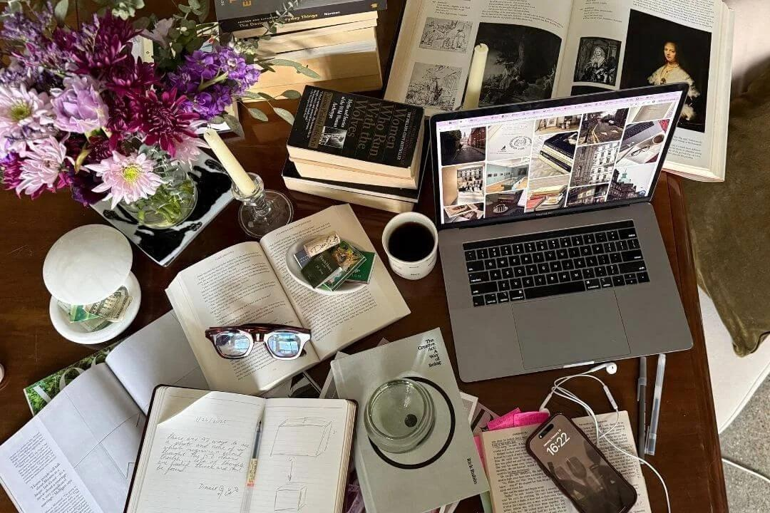 A lived-in table with books, handwritten notes, flowers, coffee, a laptop, and a phone